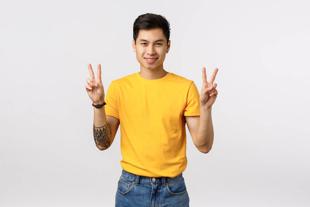 Stylish good-looking asian man with tattoos in yellow t-shirt, showing peace signs, victory gesture, smiling joyfully, send positive vibes, having fun, standing in playful mood, white background - Photo, Image