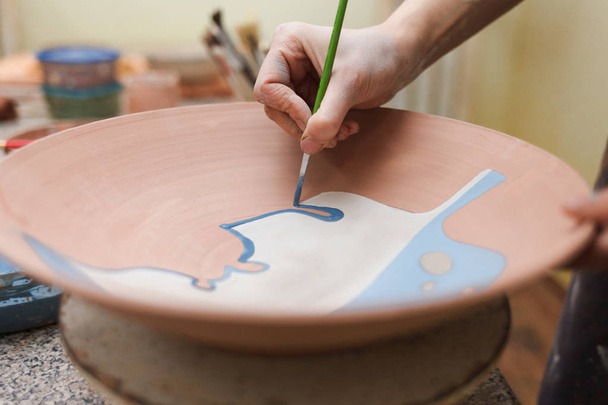 Potter žena maluje keramické desky detailní. Dívka kreslí štětcem na hliněné nádobí. Proces tvorby jílovitých výrobků. - Fotografie, Obrázek