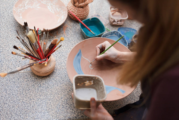 Potter woman paints a ceramic plate. Girl draws with a brush on earthenware. Process of creating clay products - Foto, Bild