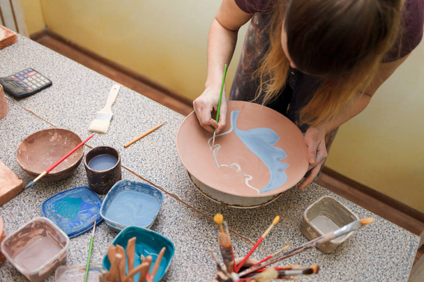 Potter denen kadın seramik tabak boyuyor. Kız toprak eşyalar üzerinde bir fırçayla resim çiziyor. Kil ürünleri oluşturma süreci - Fotoğraf, Görsel