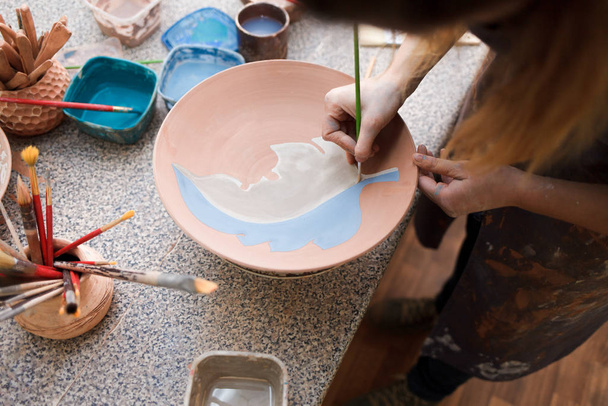 Töpferin malt einen Keramikteller. Mädchen zeichnet mit Pinsel auf Steingut. Prozess der Herstellung von Tonprodukten - Foto, Bild