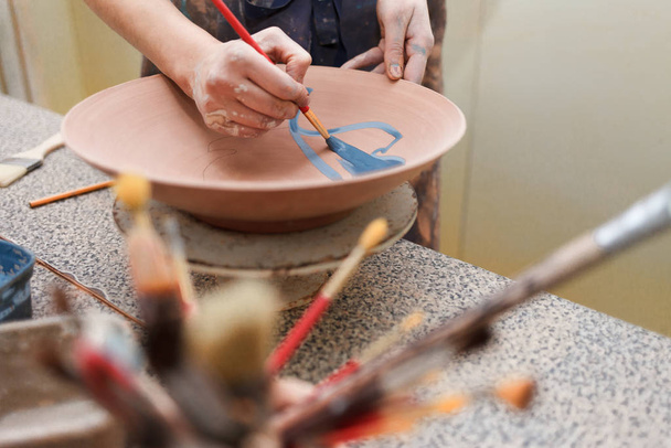 Potter woman paints a ceramic plate. Girl draws with a brush on earthenware. Process of creating clay products - 写真・画像