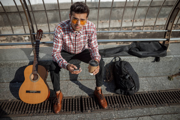 Young guy looking at camera while sitting with guitar neat to him - Photo, Image
