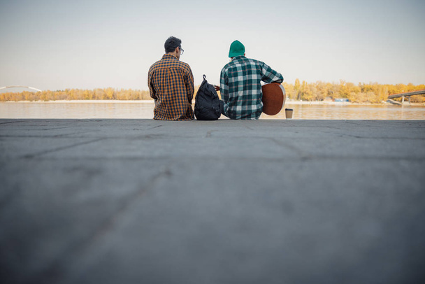 Two male friends siting with guitar near river - 写真・画像