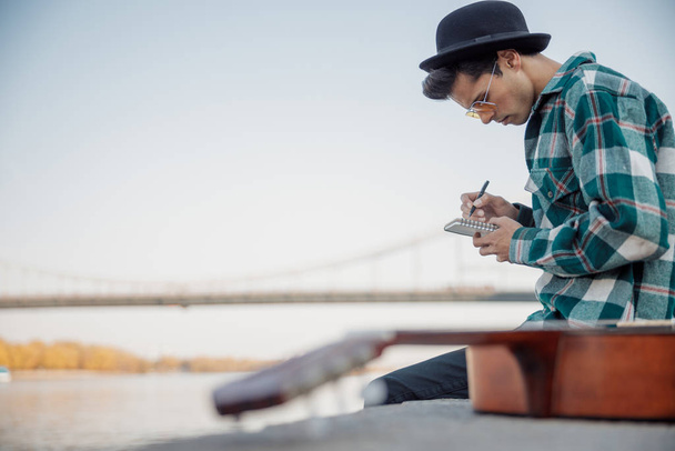 Young guy with guitar sitting near river - Φωτογραφία, εικόνα