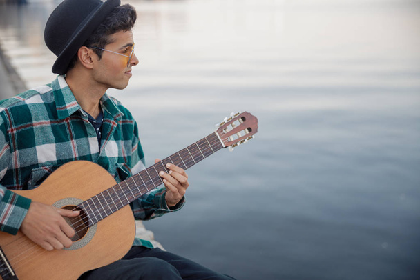Smiling guy with guitar sitting near river - Foto, Imagen