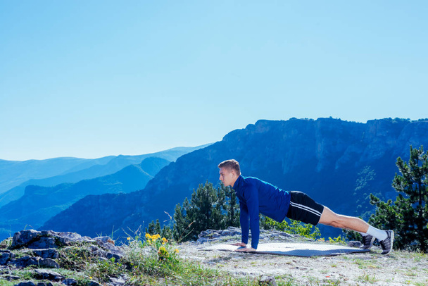 Sterk knappe blonde atleet doet push-ups op de top van en berg terwijl u geniet van het geweldige uitzicht op het meer in de vallei. - Foto, afbeelding