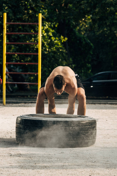 Sportovní muž bez trička v černém sportovním oblečení tlačí velký pneumatiky trénink v ulici tělocvična - Fotografie, Obrázek