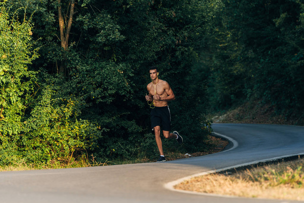 Άνδρας δρομέας χωρίς πουκάμισο με ακουστικά. Άνθρωπος τρέχει στο ηλιοβασίλεμα στον ορεινό δρόμο - Φωτογραφία, εικόνα