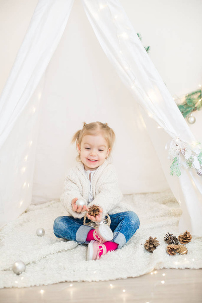Malá roztomilá holčička v bílém teplém pleteném svetru u dětské lóže zdobené věncem a jehlicemi, hračkami a mandarinkami. vánoční nálada - Fotografie, Obrázek