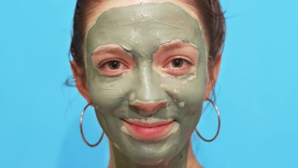 Makró portré egy tisztító agyag maszkos nőről, aki boldogan mosolyog a kék háttérben. Női arc barna szemek alkalmazása természetes kozmetikumok közelről. Arcápolás koncepció egészségügyi bőrápolás - Felvétel, videó