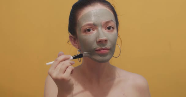 化粧ブラシを使用して顔マスクを適用パンと自然なブルネットは黄色の背景をモックアップします。屋内スローモーションコピースペースをスキンケアを取って若い女性のフロント肖像画。女性美容室 - 映像、動画