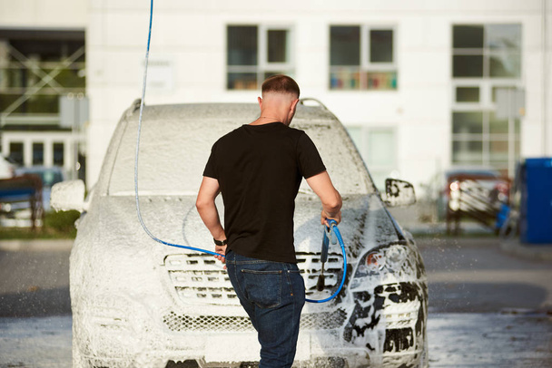 На задньому плані - спортивний вродливий чоловік, що тримає шланг з милом і миє свій автомобіль на вулиці, біла піна на чорній машині. - Фото, зображення