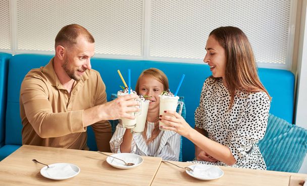 glückliche Familie - Mama, Papa und Tochter sitzen im örtlichen Café auf einem blauen Sofa und trinken Getränke, klimpern mit Milchshakes. Teller und Cocktaillöffel auf dem Tisch - Foto, Bild