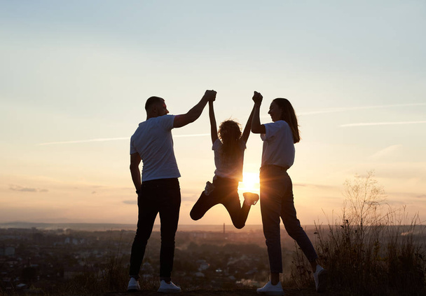 Silhouette of father, mother and daughter having fun outside the city on the hill on the sunset with a beautiful city view, man and woman holding girl by hands and she is jumping, back view - 写真・画像
