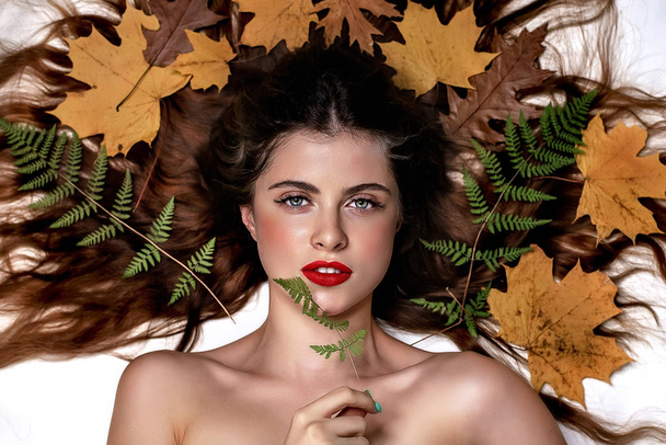 Portrait of a young brunette woman on a white background with a green fern and yellow leaves around her head - Photo, Image