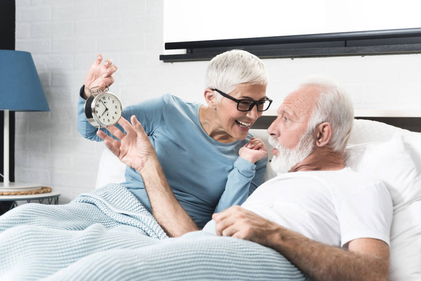 Early happy morning in modern beautiful bedroom  for senior aged couple with white hair smile. Senior woman holds an alarm clock and wants to wake her sleeping husband.  Surprised face on the man. The concept time is precious - Fotoğraf, Görsel