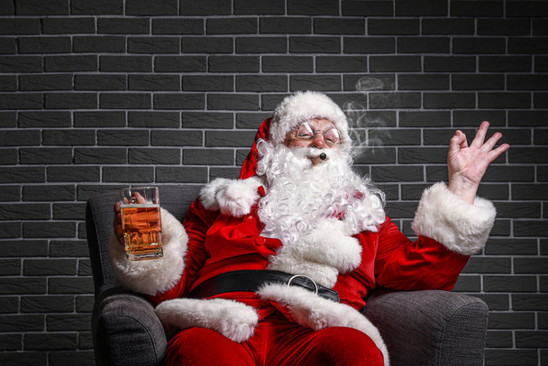 Funny drunk Santa Claus showing OK while sitting in armchair against brick background - Fotó, kép