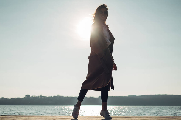 Krásná mladá stylová dívka ve kabátě chůze na jarní pláži při západu slunce - Fotografie, Obrázek