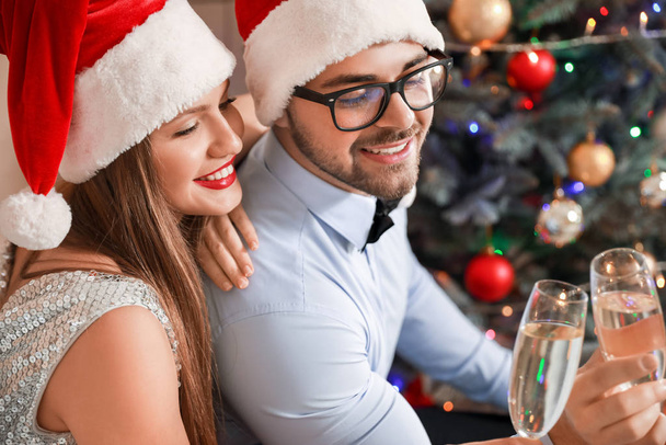 Szczęśliwa młoda para w kapeluszach Świętego Mikołaja i kieliszkach szampana w domu - Zdjęcie, obraz