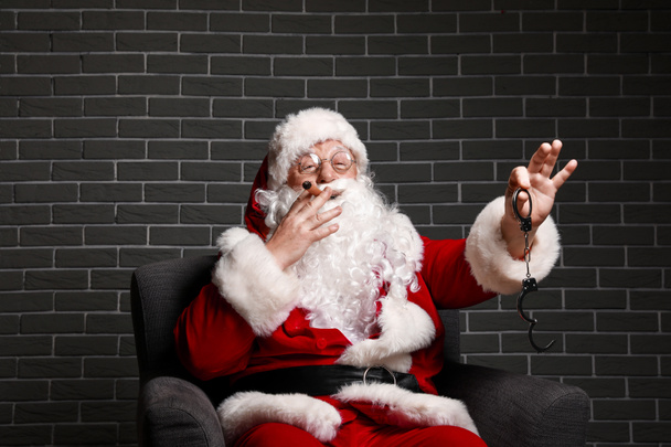 Αστείος μεθυσμένος Άγιος Βασίλης με χειροπέδες κάθεται στην πολυθρόνα με φόντο τούβλο - Φωτογραφία, εικόνα