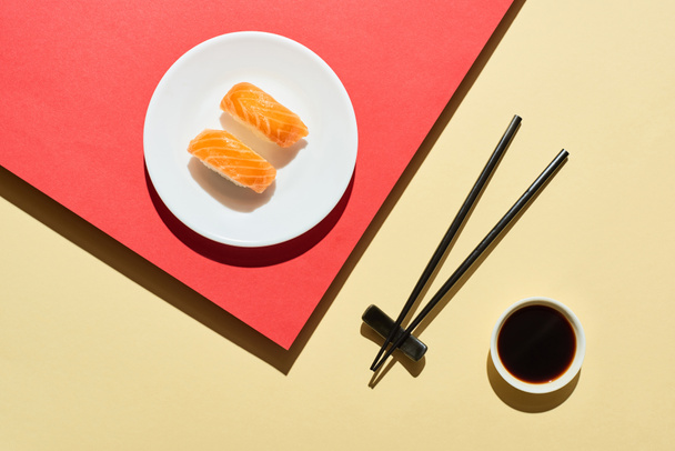 κορυφαία άποψη των φρέσκων nigiri με σολομό κοντά σάλτσα σόγιας και chopsticks σε κόκκινο και μπεζ επιφάνεια - Φωτογραφία, εικόνα