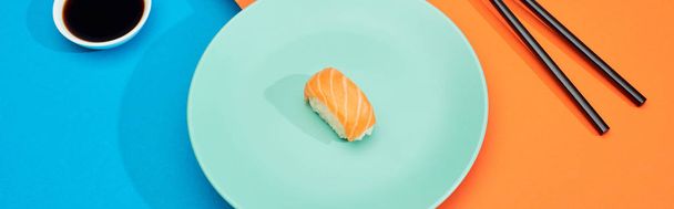 fresh nigiri with salmon near soy sauce and chopsticks on blue and orange surface, panoramic shot - 写真・画像