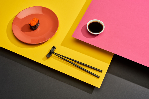 verse maki met rode kaviaar op bord bij eetstokjes en sojasaus op roze, gele en zwarte ondergrond - Foto, afbeelding