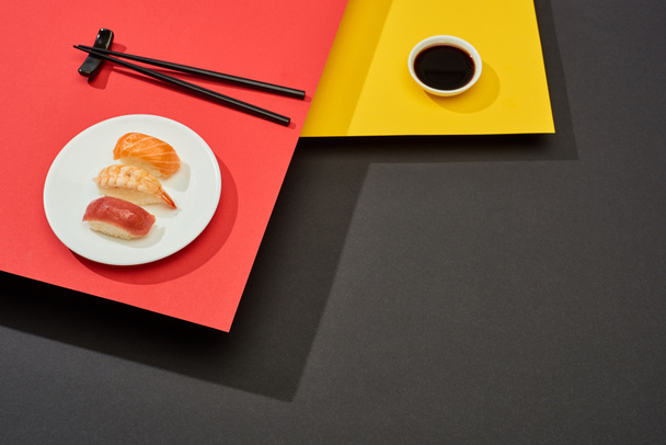 свежий нигири с лосося, креветок и тунца рядом соевый соус и палочки для еды на красной, желтой и черной поверхности
 - Фото, изображение