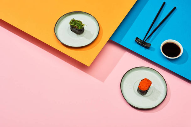 fresh nigiri with seaweed and red caviar near soy sauce and chopsticks on blue, pink, orange surface - Photo, Image