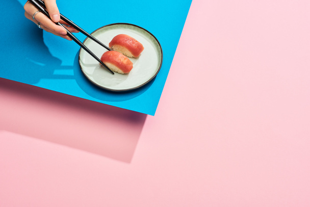 vista parcial de la mujer comiendo nigiri fresco con atún con palillos sobre fondo azul, rosa
 - Foto, Imagen