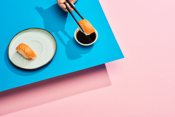 cropped view of woman putting fresh nigiri with salmon into soy sauce on blue, pink background - Photo, Image