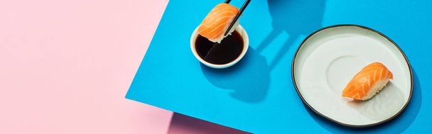 fresh nigiri with salmon putting in soy sauce on blue, pink background, panoramic shot - Photo, Image