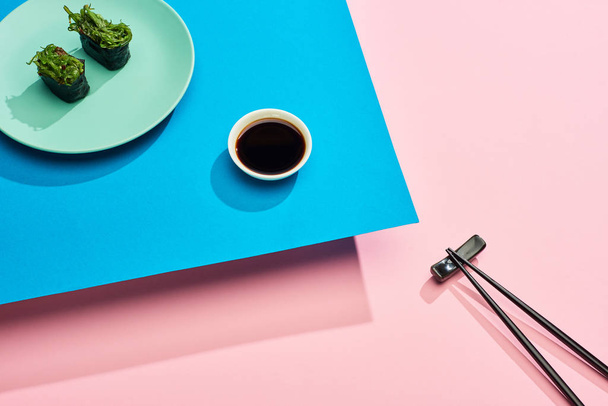 свежие нигири с водорослями рядом с соевым соусом и палочками для еды на голубом, розовом фоне
 - Фото, изображение