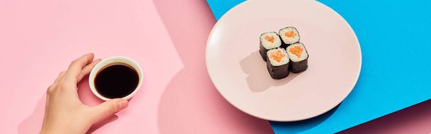 cropped view of woman holding soy sauce near fresh maki with salmon on blue, pink background, panoramic shot - Photo, Image