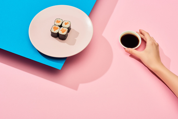 cropped view of woman holding soy sauce near fresh maki with salmon on blue, pink background - Fotó, kép