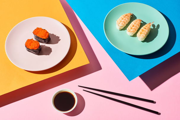 πάνω όψη φρέσκων nigiri με γαρίδες και κόκκινο χαβιάρι κοντά σε σάλτσα σόγιας και chopsticks σε μπλε, ροζ, πορτοκαλί φόντο - Φωτογραφία, εικόνα