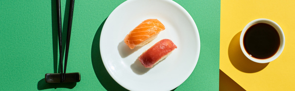 top view of fresh nigiri with salmon and tuna near soy sauce and chopsticks on green and yellow surface, panoramic shot - Fotografie, Obrázek