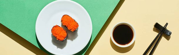 top view of fresh nigiri with red caviar near soy sauce and chopsticks on green and yellow surface, panoramic shot - 写真・画像