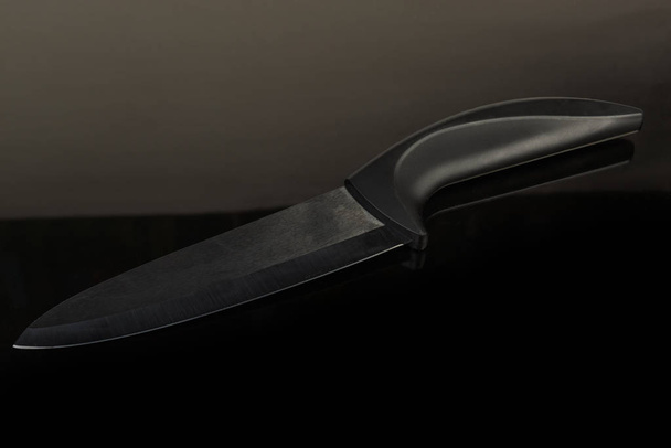 Кухонный керамический нож с черным лезвием на темной отражающей поверхности
 - Фото, изображение