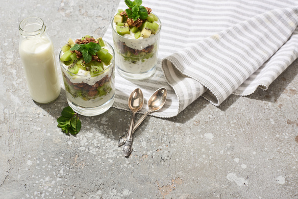 granola fresca con kiwi y yogur en superficie de hormigón gris con servilleta rayada, botella de leche y cucharas
 - Foto, Imagen