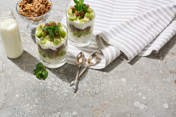 granola fresca con kiwi y yogur en superficie de hormigón gris con servilleta rayada, botella de leche, nueces y cucharas
 - Foto, Imagen