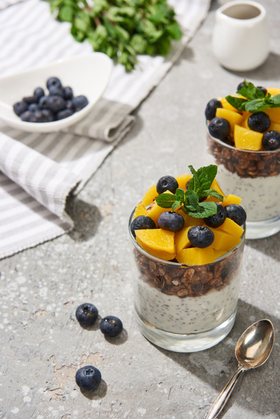 selective focus of fresh granola with canned peach, blueberries and chia seeds on grey concrete surface with spoon and napkin - Foto, Imagen