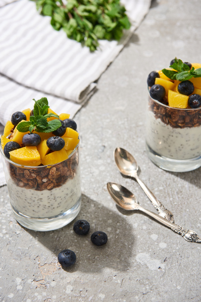 selective focus of fresh granola with canned peach, blueberries and chia seeds on grey concrete surface with spoons, mint and napkin - Φωτογραφία, εικόνα