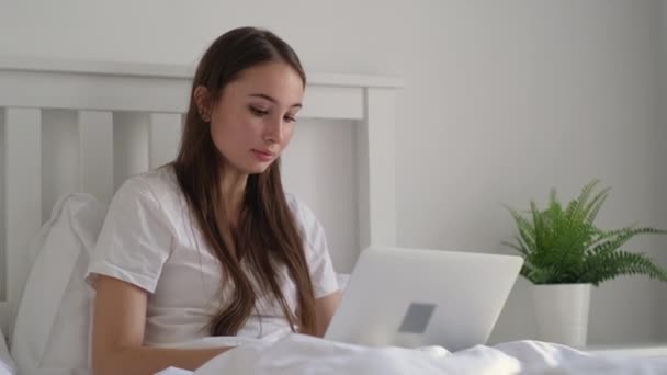 Girl chatting online on laptop staying in bed - Кадры, видео