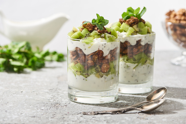 selective focus of fresh granola with kiwi and yogurt near spoons on concrete surface isolated on grey - Foto, Imagen
