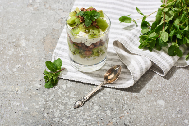 granola fresca con kiwi y yogur en superficie de hormigón gris con servilleta a rayas, cuchara y menta
 - Foto, Imagen