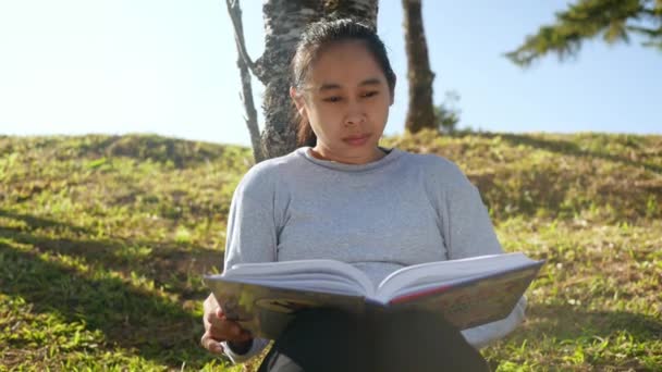 Asian relaxed young woman sitting under a tree in spring garden and reading a book with sun shining. - Metraje, vídeo
