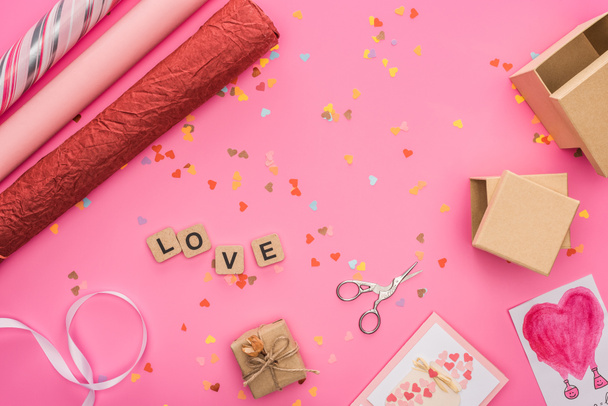 top view of valentines confetti, scissors, wrapping paper, gift boxes, greeting cards and love lettering on wooden cubes on pink background - Photo, image