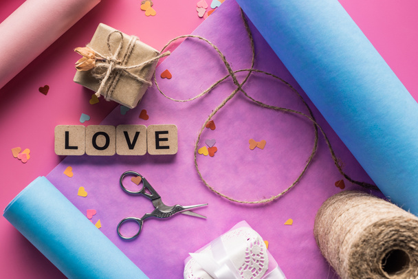 top view of valentines decoration, scissors, wrapping paper, twine, gift boxes and love lettering on wooden cubes on pink background - Fotografie, Obrázek